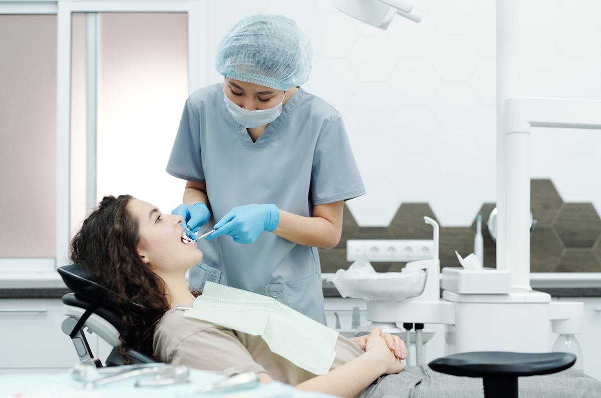 Woman in Blue Scrub Suit Standing Beside Woman in Brown Shirt Lying on Dental Chair