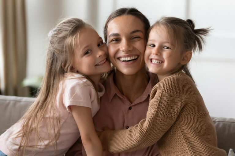 Beautiful female family young smiling single mother and cute little children daughters embracing looking at camera, funny small kids girls hugging happy mum bonding sit on sofa together, portrait
