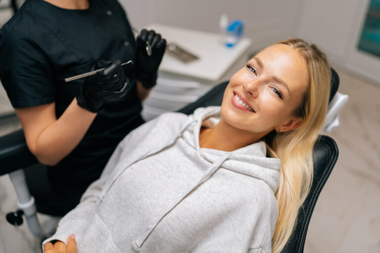 Portrait of charming young woman patient with perfect white teeth and smile satisfied after dental treatment lying on dental chair in dentistry clinic. Female dentist in gloves examining teeth.